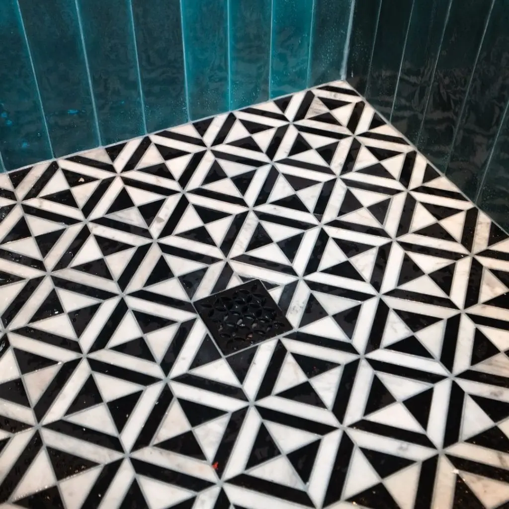 Close-up of a patterned black, white, and gray mosaic tile shower floor with a decorative square drain in the center.