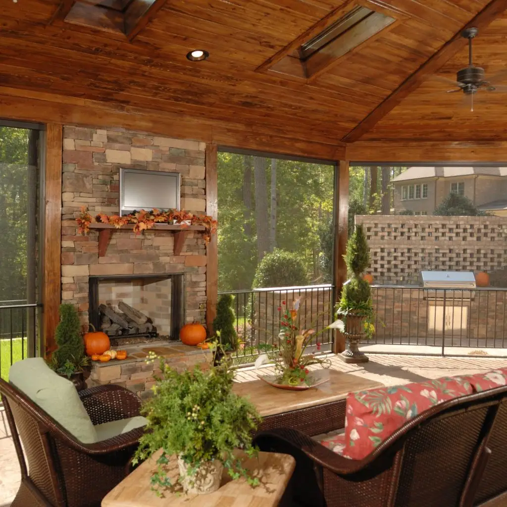 Cozy screened-in porch featuring a rustic stone fireplace with seasonal decor, warm wood ceiling with skylights, comfortable wicker furniture, and lush greenery creating a relaxing outdoor living space.