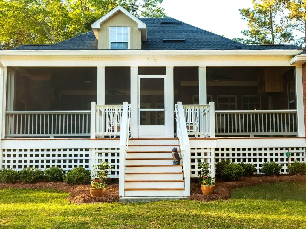 Front view of a charming Southern-style screened-in porch with white railings, a central staircase, decorative lattice skirting, and rocking chairs, framed by landscaped flower beds and lush green lawn.