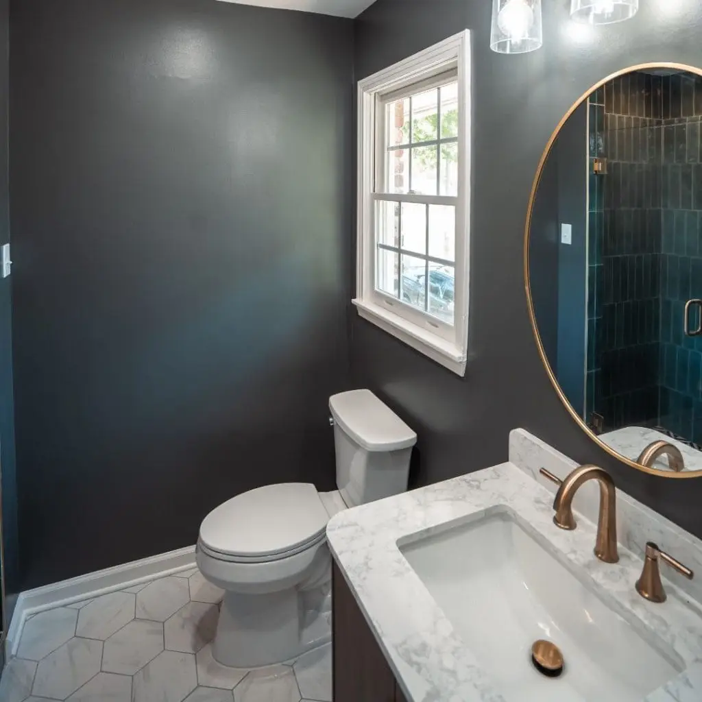 Modern bathroom with a dark gray wall, white toilet, hexagon floor tiles, marble countertop vanity with brushed gold fixtures, and a round mirror.