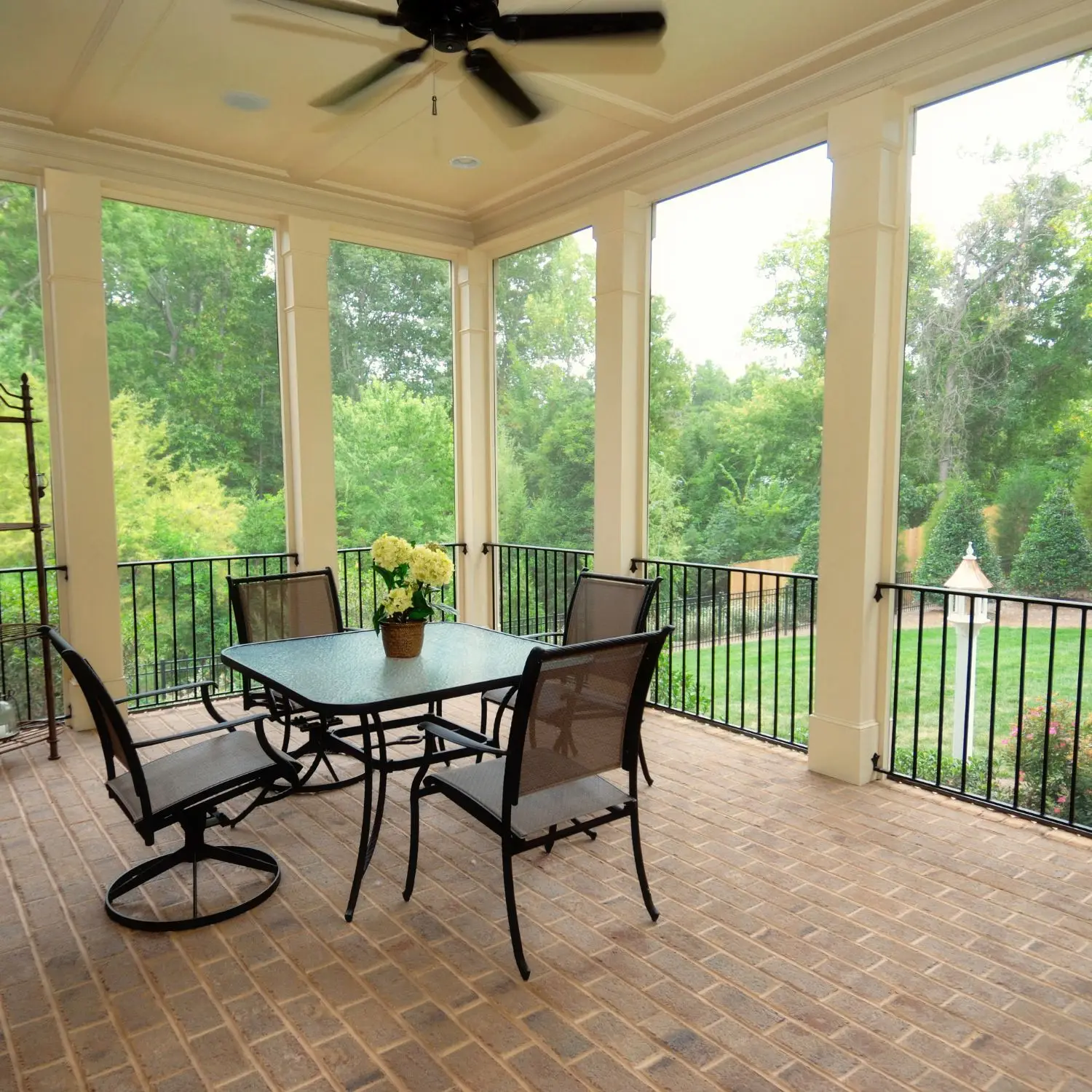 Spacious screened-in porch with tall, elegant columns, brick flooring, black railing, ceiling fan, and comfortable outdoor dining set overlooking a lush, green backyard.