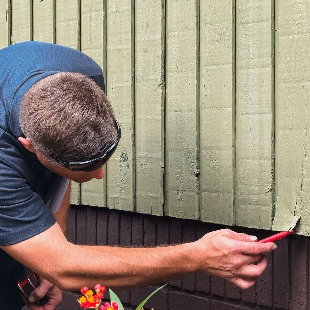 Inspector examining cracked and peeling green siding at the base of a home for signs of moisture damage.